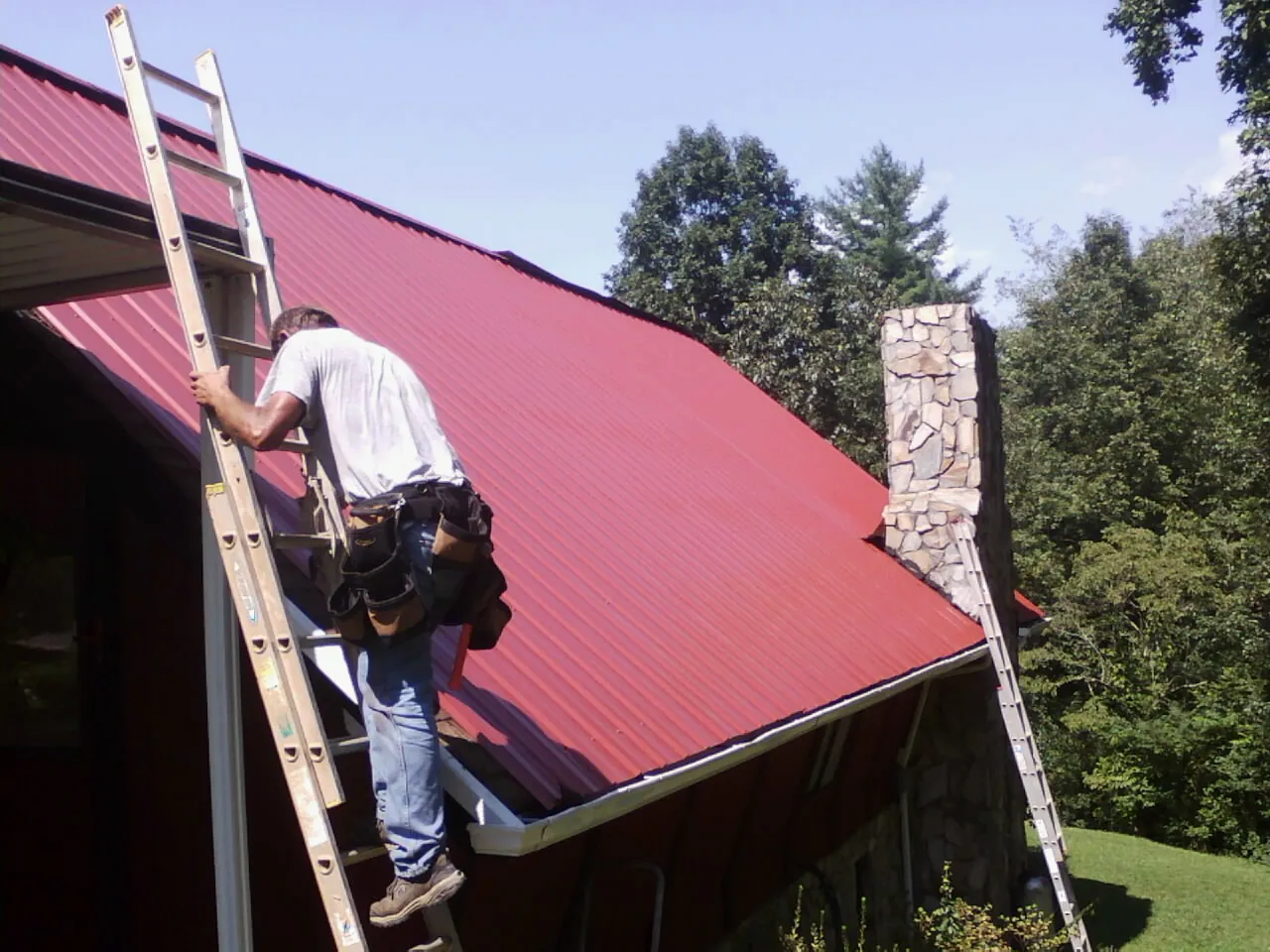 Roofer on a ladder beside a red metal roof and stone chimney — Soffit & Fascia Repair by Roofing Brothers in Red Cliff